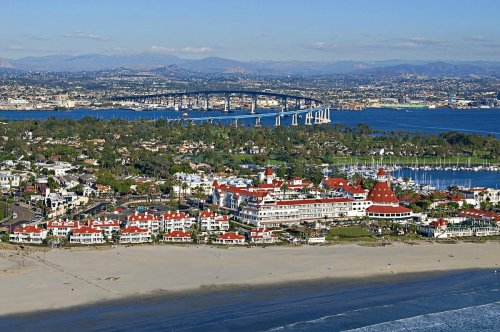 Aerial view of a coastal resort with red-roofed buildings, beach, and a bridge over blue water in the background.