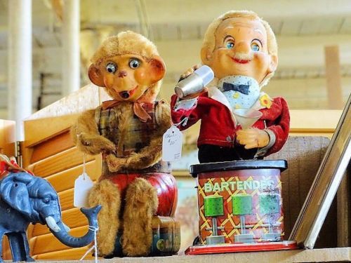 Two vintage dolls: a monkey in a plaid shirt and a smiling bartender holding a shaker, displayed on a shelf.