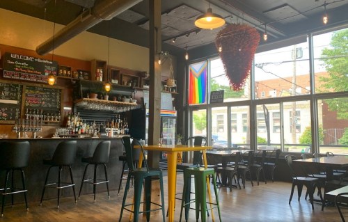 A cozy bar interior with a colorful pride flag, wooden tables, and a well-stocked bar in a bright, welcoming space.