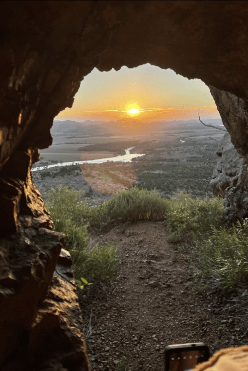 View of a sunset through a rocky cave opening, with a river and lush landscape in the background.