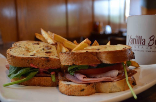 A hearty sandwich with layers of meat, spinach, and tomato, served with golden fries on a white plate.