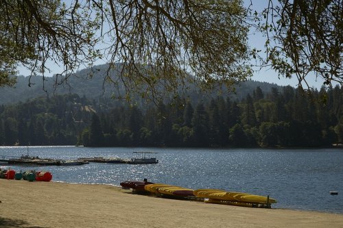 A serene lake scene with colorful kayaks on the shore, surrounded by trees and mountains under a clear blue sky.