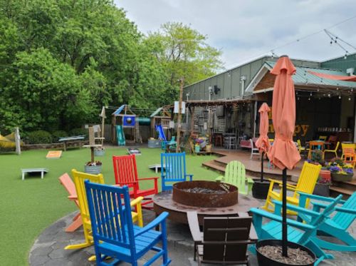 Colorful chairs surround a fire pit in a vibrant outdoor space with greenery and a rustic building in the background.