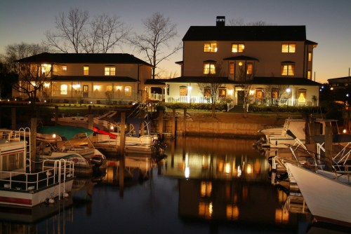 A serene marina at dusk, featuring illuminated buildings and boats reflecting in calm water.