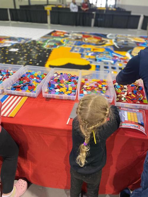 A child with braided hair explores colorful LEGO bricks at a table, with a large LEGO mosaic in the background.