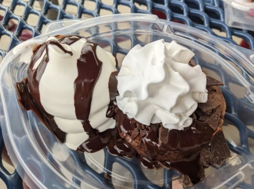 A plastic container holds two scoops of ice cream, one vanilla and one chocolate, topped with whipped cream and chocolate sauce.