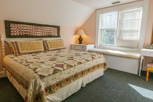 Cozy bedroom with a quilted bedspread, window seat, and soft lighting from a bedside lamp.