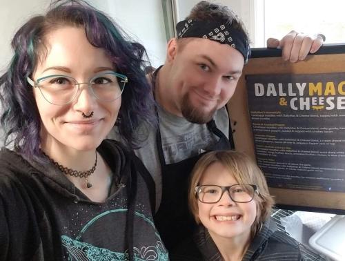 A group of three people smiling in a kitchen, holding a sign for "Dally Mac & Cheese."
