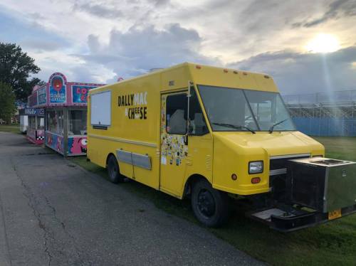 A yellow food truck named "Dally Mac Cheese" parked next to a colorful carnival stand under a cloudy sky.