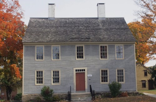 A historic two-story blue house with a red door, surrounded by autumn trees and a clear sky.