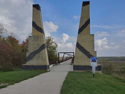 A modern bridge with tall, patterned concrete pillars, surrounded by greenery and a cloudy sky.