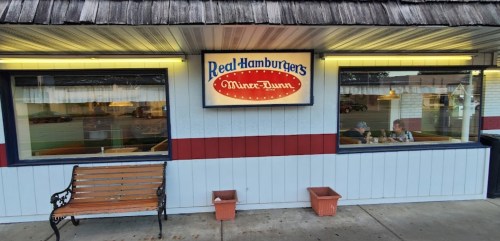 Exterior of a diner with a sign reading "Real Hamburgers" and a bench outside, featuring large windows and a cozy atmosphere.