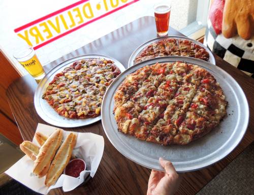 A hand holds a large pizza on a metal tray, with two more pizzas and breadsticks on the table, alongside two beers.