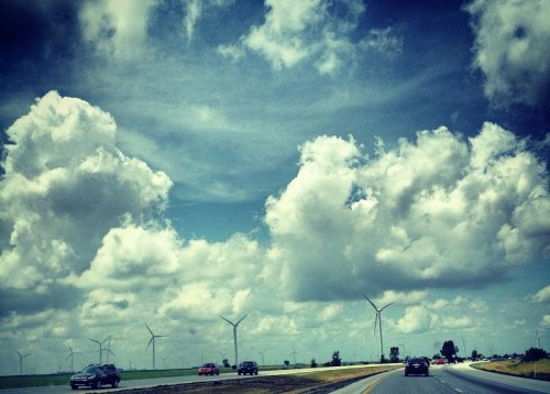 A scenic highway with cars driving, surrounded by green fields and wind turbines under a cloudy sky.