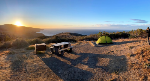 A scenic campsite at sunset, featuring a green tent, picnic table, and ocean view in the background.