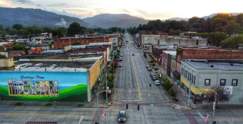 Aerial view of a small town street with a mural, mountains in the background, and cars lining the road at sunset.