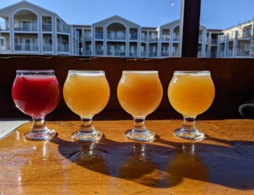 Four small glasses of colorful drinks on a wooden table, with a sunny view of a building in the background.