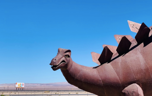 A large, rusty dinosaur sculpture against a clear blue sky, with a billboard visible in the background.