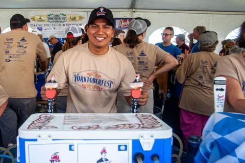 A smiling man stands behind a beer tap at a festival, surrounded by people in casual attire.