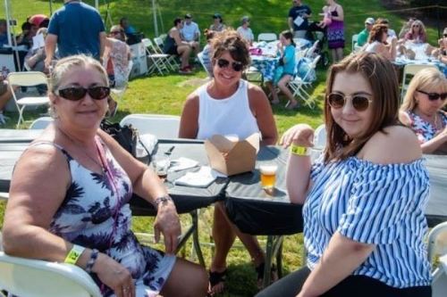 Three women smiling at a table outdoors, enjoying a sunny day at a festival with people in the background.