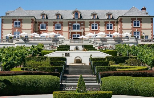 A grand estate with a red and cream facade, surrounded by manicured gardens and vineyards under a clear blue sky.