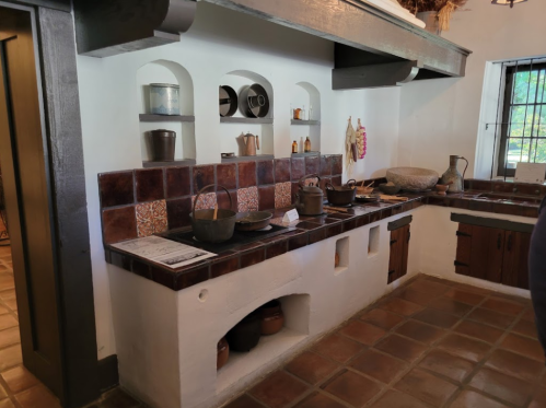 A rustic kitchen with a tiled counter, various pots, and traditional cooking tools displayed on shelves.