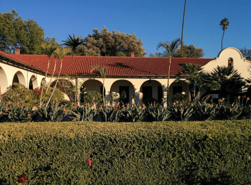 A charming building with a red tile roof, surrounded by lush greenery and palm trees under a clear blue sky.