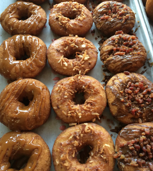 A tray of assorted donuts, featuring caramel, coconut, and bacon toppings, arranged in two rows.