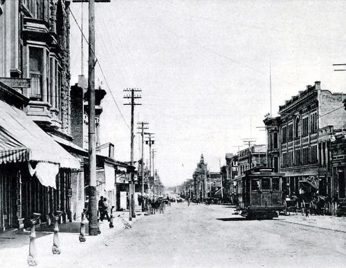 Black and white historical street scene with horse-drawn carriages, buildings, and power lines.