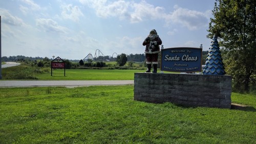 A welcome sign for Santa Claus, Indiana, featuring a Santa statue and a blue Christmas tree, with a roller coaster in the background.