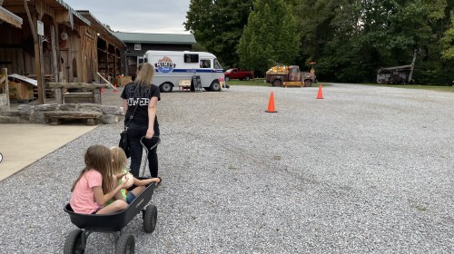 A person pulls a wagon with two children through a gravel area near a food truck and orange cones.