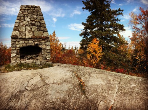 A stone fireplace on a rocky surface, surrounded by colorful autumn trees under a blue sky.