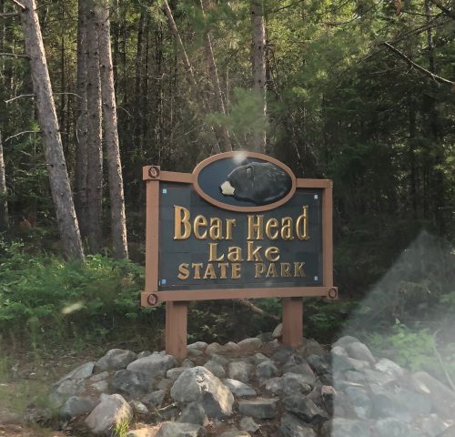 Sign for Bear Head Lake State Park, featuring a bear graphic, surrounded by trees and rocky terrain.