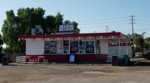 A small fast-food restaurant with a red and white exterior, outdoor seating, and a sign displaying menu items.