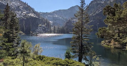 A serene lake surrounded by mountains and trees, reflecting sunlight on the water's surface.