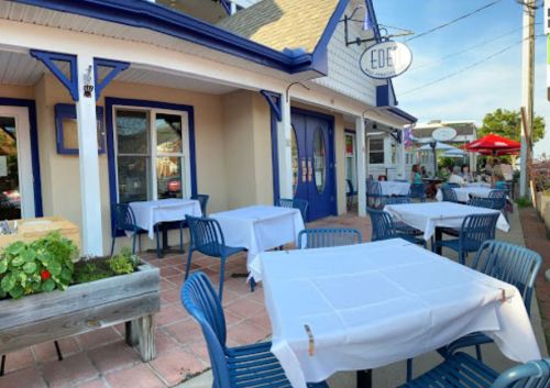 Outdoor dining area of a restaurant with white tablecloths, blue chairs, and potted plants, under a clear sky.