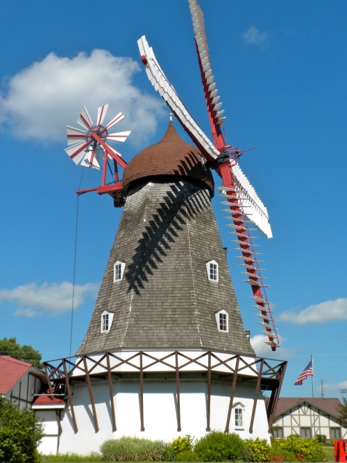 A traditional windmill with red blades, set against a blue sky with fluffy clouds and greenery in the foreground.
