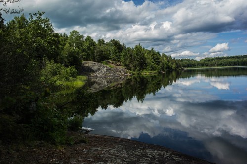 A serene lake surrounded by lush greenery, reflecting clouds and trees in calm waters under a partly cloudy sky.