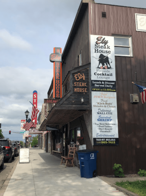 Exterior of Ely Steak House with signage, flags, and a cloudy sky in the background.