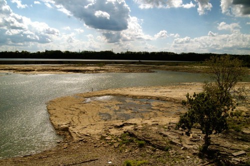 A serene landscape featuring a calm river, rocky shore, and scattered clouds in a blue sky.