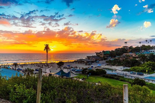 Sunset over a beach with palm trees, colorful clouds, and people enjoying the shoreline and surrounding park.