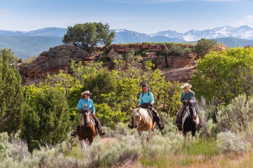 Three riders on horseback traverse a scenic landscape with mountains in the background and lush greenery around them.