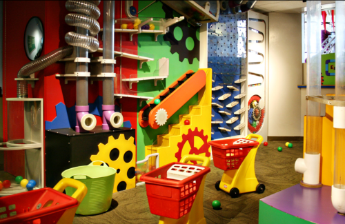 Colorful play area with interactive toys, gears, and slides, featuring red toy carts and various playful structures.