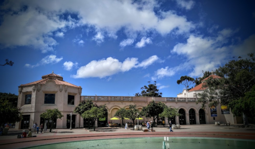 A sunny day at a plaza featuring a historic building, trees, and a fountain under a blue sky with fluffy clouds.