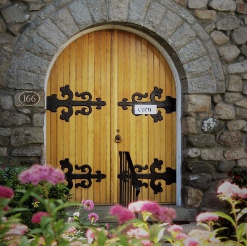 A wooden arched door with decorative metal hinges, set in a stone wall, surrounded by colorful flowers.