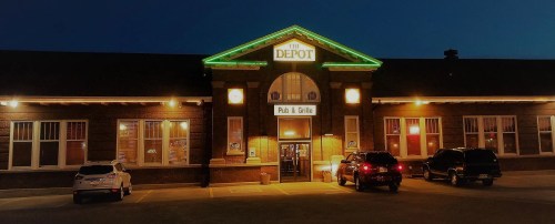 The Depot Pub & Grille illuminated at night, featuring a brick facade and green lights. Cars parked in front.
