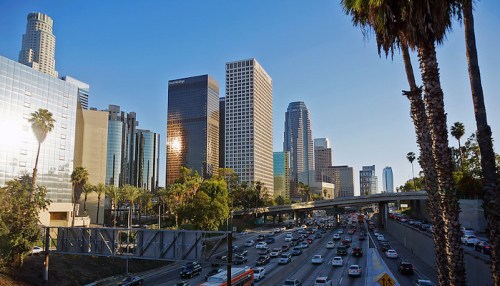 A bustling Los Angeles skyline with tall buildings, palm trees, and a busy highway under a clear blue sky.