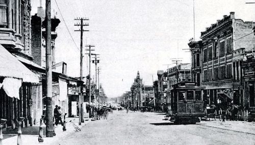 Black and white photo of a historic street with a tram, horse-drawn carriages, and early 20th-century buildings.