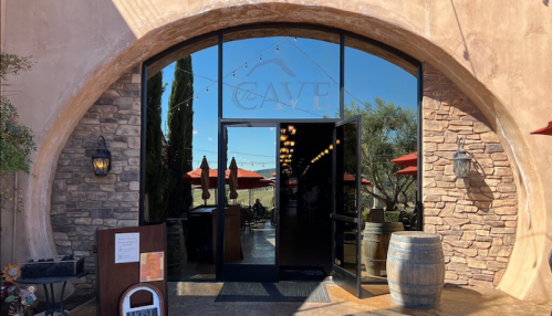Entrance to The Ave, featuring large glass doors, stone walls, and outdoor seating with red umbrellas.