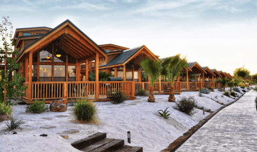 A row of wooden cabins with porches, surrounded by palm trees and landscaped paths under a clear sky.